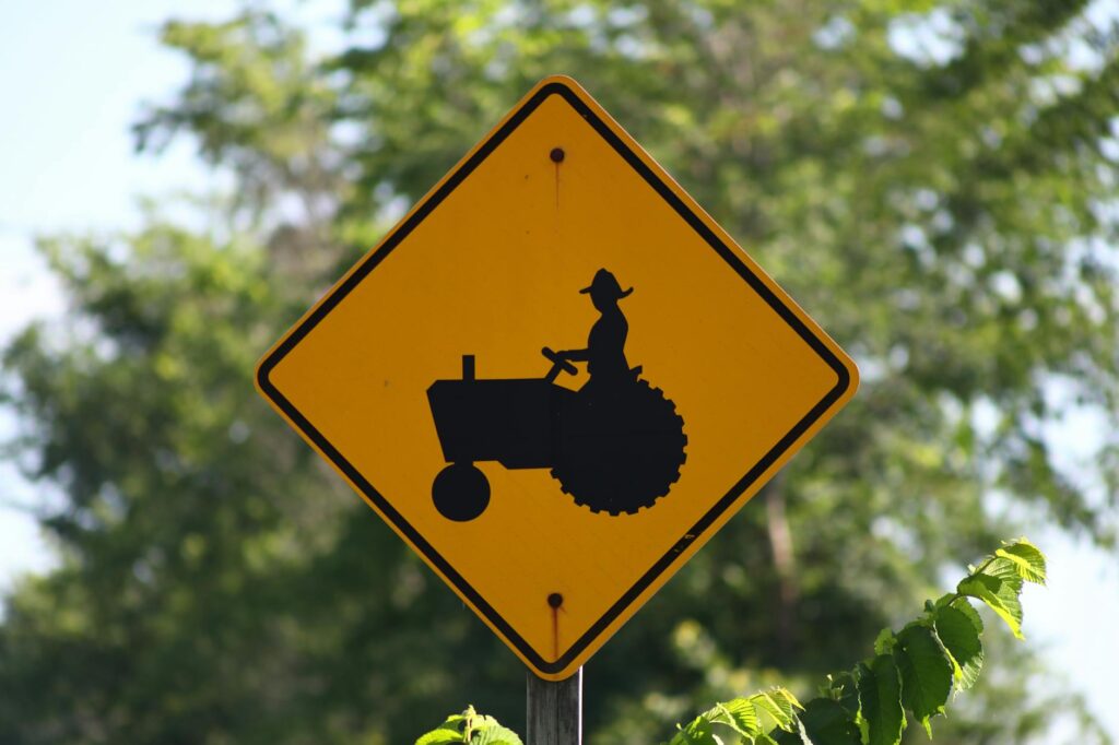 Close-up of a yellow tractor crossing sign on a rural road, set against a lush green backdrop.