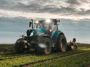 Big tractor with headlights and plow riding on green agricultural plantation in countryside on farmland against cloudy sky during harvest season